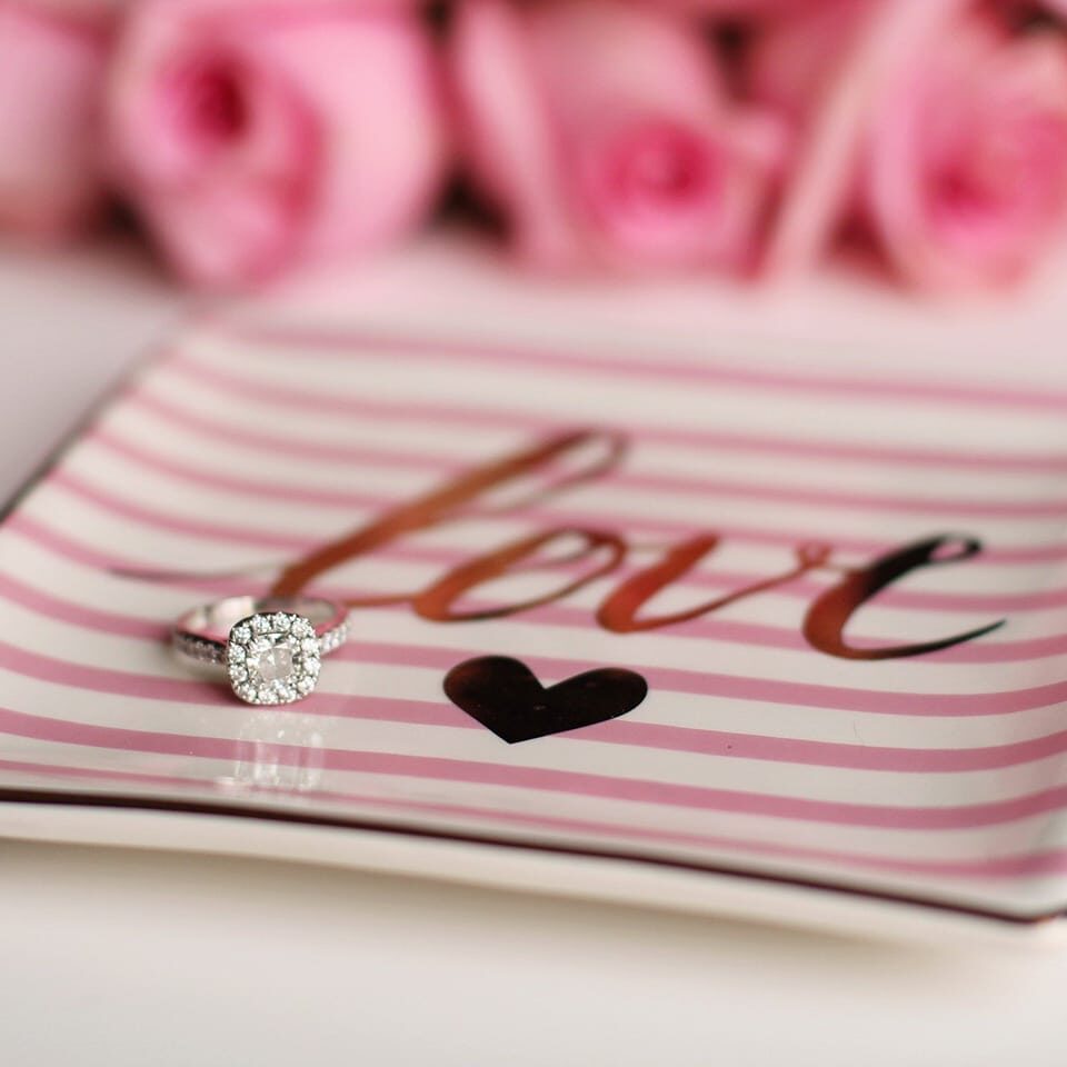 A diamond ring on a pink-striped plate with roses in the background.